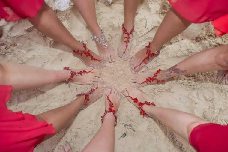 Bridal party's feet in a circle on the sand wearing red barefoot sandals